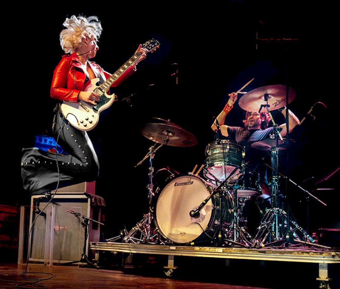 SAMANTHA FISH Paper Doll Live Photo Credit © Doug Hardesty - female guitar player in red jumping-up on stage with drummer behind
