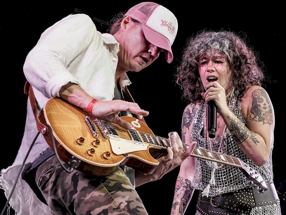 BYWATER CALL Broken Souvenirs photo credit © Paul May - female singer with tattoos on her arms singing into microphone over the neck of an electric guitar played by a man wearing a baseball cap