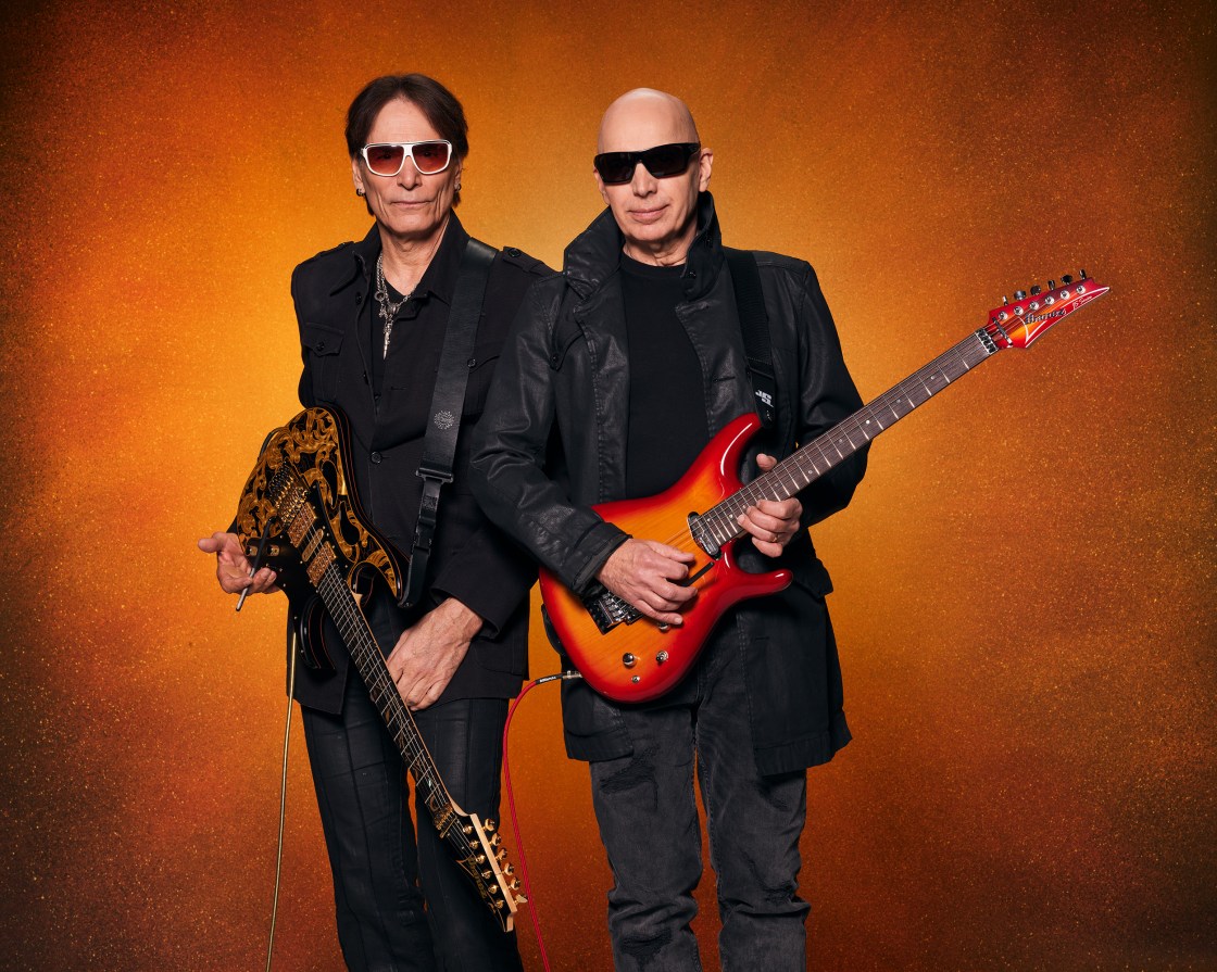 SATCHVAI Explore Sea of Emotion - Steve Vai_and Joe Satriani_ - photo by Larry DiMarzio - wto white men holding guitars against an orange backdrop